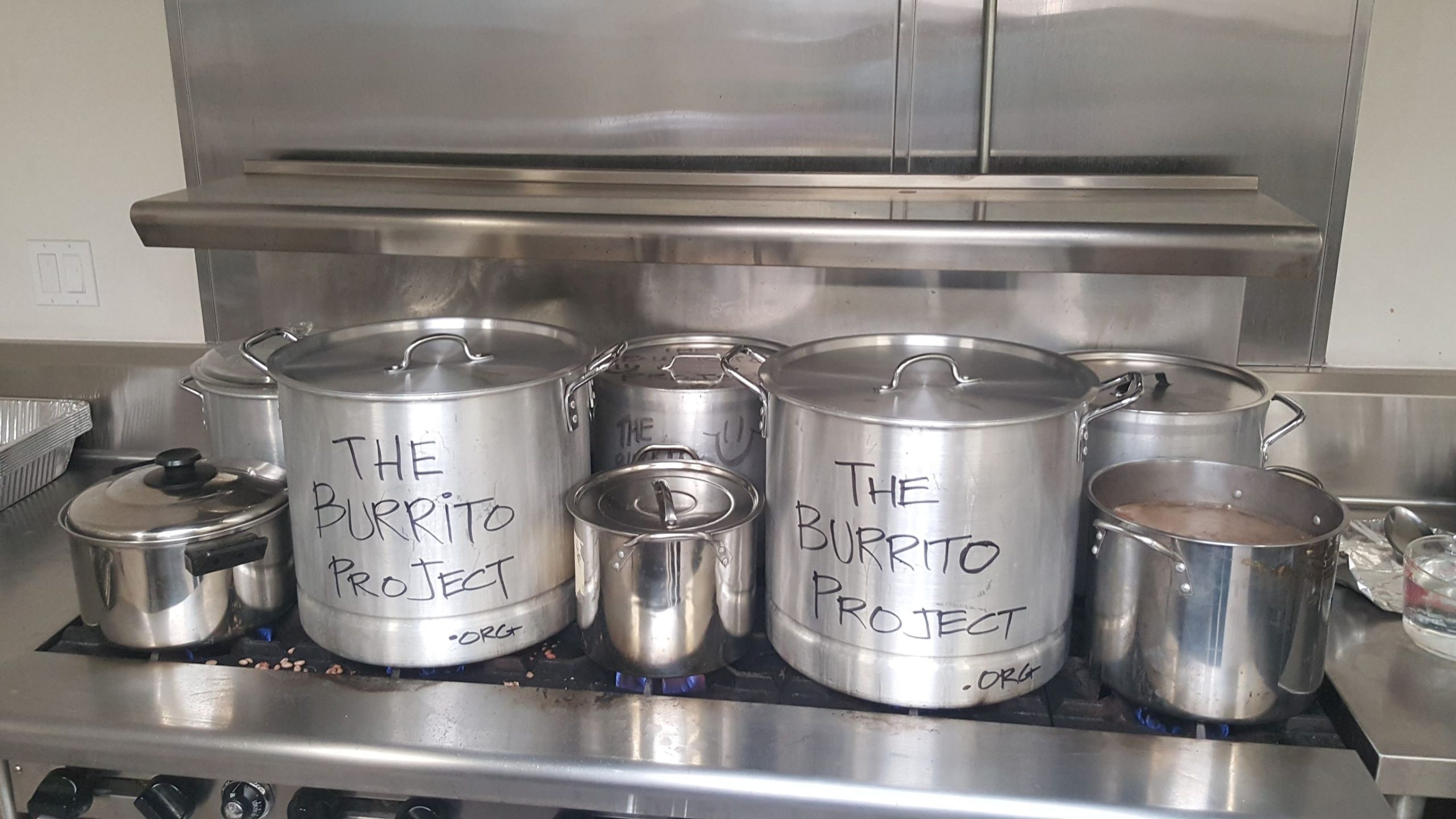 Industrial pots labeled The Burrito Project simmering on a commercial stove