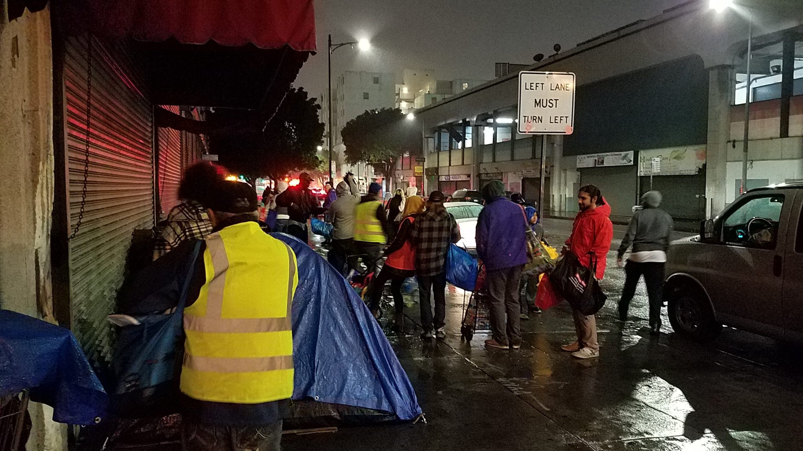 Volunteers in yellow vests delivering meals on a rainy downtown LA street at night